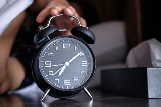A clock on a bed side table with someone reaching for it.