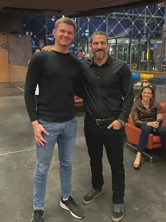 Two men smiling and posing together in a modern lounge area with a woman sitting behind them.