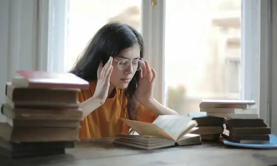 Stressed woman with glasses rubbing her temples while studying with stacks of books.